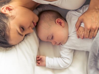 Healthy mom naps with sleeping baby in white long sleeved onesie holding baby's hand.